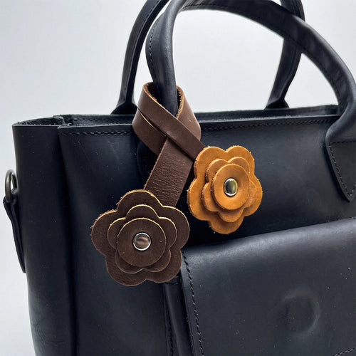 A close-up of a black leather handbag with two decorative flower-shaped leather charms attached to the handle. One charm is medium brown and the other is mustard yellow, both with metal snap buttons.