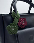 A close-up of a black leather handbag featuring two decorative flower-shaped leather charms attached to the handle. One charm is olive green with a silver-tone center, and the other is burgundy with a silver-tone center.