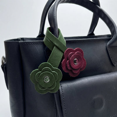 A close-up of a black leather handbag featuring two decorative flower-shaped leather charms attached to the handle. One charm is olive green with a silver-tone center, and the other is burgundy with a silver-tone center.