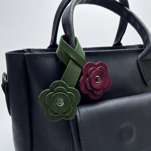 A close-up of a black leather handbag featuring two decorative flower-shaped leather charms attached to the handle. One charm is olive green with a silver-tone center, and the other is burgundy with a silver-tone center.
