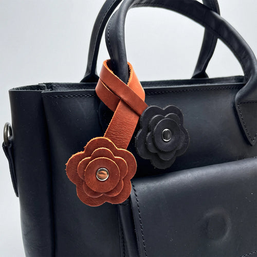 A close-up of a black leather handbag featuring two decorative flower-shaped leather charms hanging from the handle. One charm is a warm brown with a silver-tone center, and the other is black with a matching center.
