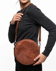 A woman wearing a dark gray long-sleeve top carries a round brown leather crossbody bag with a geometric stitched pattern and a brown shoulder strap against a white background.