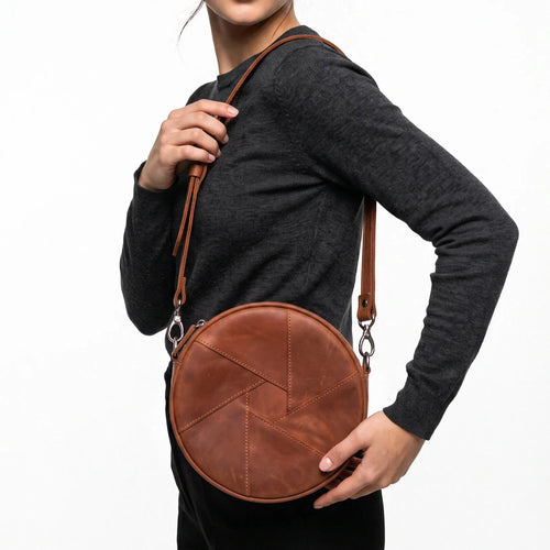A woman wearing a dark gray long-sleeve top carries a round brown leather crossbody bag with a geometric stitched pattern and a brown shoulder strap against a white background.