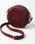 A round burgundy leather crossbody bag with a geometric stitched pattern, a matching adjustable leather strap with a silver buckle, and silver hardware, shown on a light gray background.