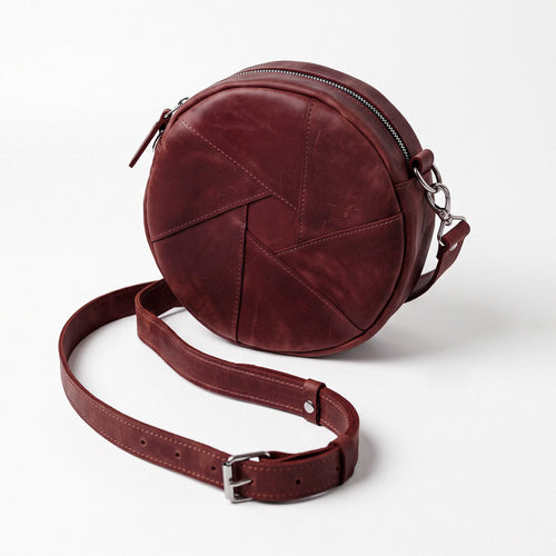 A round burgundy leather crossbody bag with a geometric stitched pattern, a matching adjustable leather strap with a silver buckle, and silver hardware, shown on a light gray background.