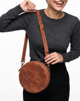 A woman wearing a dark gray long-sleeve top and black pants carries a round brown leather crossbody bag with a matching adjustable strap against a plain white background.