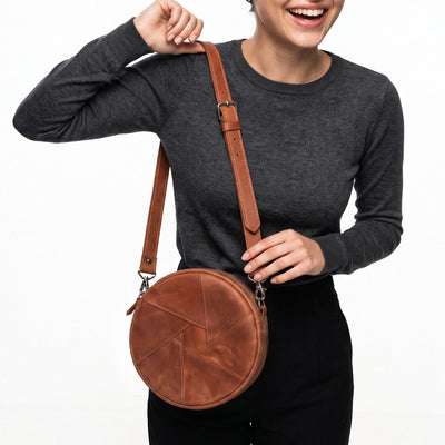 A woman wearing a dark gray long-sleeve top and black pants carries a round brown leather crossbody bag with a matching adjustable strap against a plain white background.