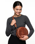 A woman wearing a dark gray long-sleeve shirt and black pants holds a brown round leather crossbody bag with a matching strap against a plain white background.