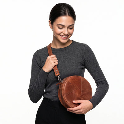 A woman wearing a dark gray long-sleeve shirt and black pants holds a brown round leather crossbody bag with a matching strap against a plain white background.