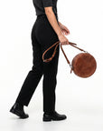 A person wearing black trousers and black boots carries a round brown leather shoulder bag with a long strap against a plain white studio background.