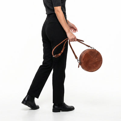 A person wearing black trousers and black boots carries a round brown leather shoulder bag with a long strap against a plain white studio background.