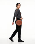 A woman in a dark gray long-sleeve top, black pants, and black ankle boots is shown from the side/back profile, carrying a round brown leather crossbody bag with a thin brown strap against a plain white studio background.