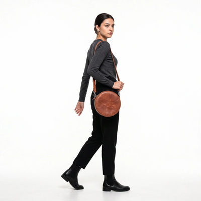 A woman in a dark gray long-sleeve top, black pants, and black ankle boots is shown from the side/back profile, carrying a round brown leather crossbody bag with a thin brown strap against a plain white studio background.