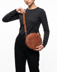A model wearing a dark gray long-sleeve top and black pants carries a round brown leather crossbody bag with a matching shoulder strap against a plain white background.