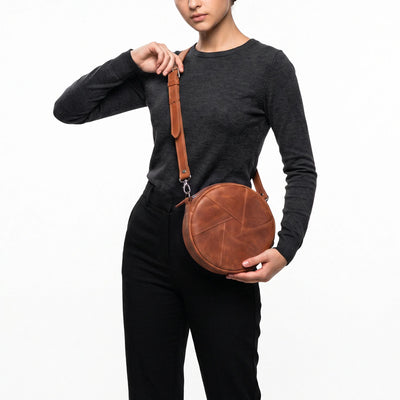 A model wearing a dark gray long-sleeve top and black pants carries a round brown leather crossbody bag with a matching shoulder strap against a plain white background.