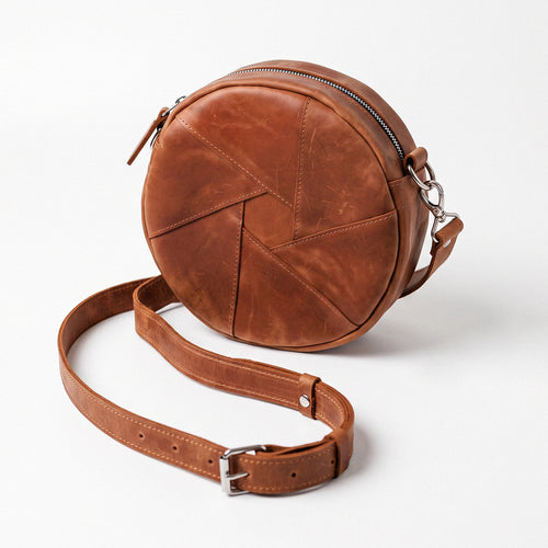 A brown leather crossbody bag with a circular body, geometric stitched panel design, and a detachable adjustable strap, shown on a light gray background.