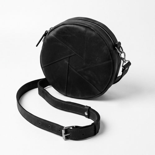 A black round leather crossbody bag with geometric stitched paneling and a detachable shoulder strap, photographed on a clean white background.