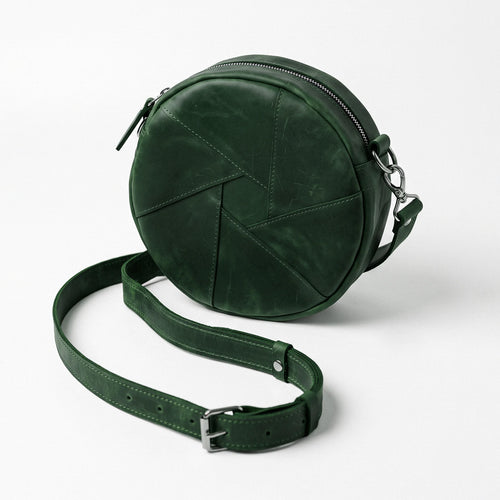 A dark green circular crossbody bag made of textured leather with geometric stitched paneling, shown with its matching adjustable shoulder strap on a light gray background.