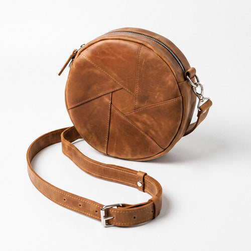 A round brown leather crossbody bag with a geometric stitched pattern, a top zipper, and a matching adjustable leather strap with a silver buckle, shown on a light gray background.
