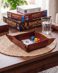 A brown leather game tray holding cards and dice, placed on a woven placemat on a wooden table. In the background, stacked BoardCARTY board games, a glass of water, and other tabletop items are visible in a real indoor setting.