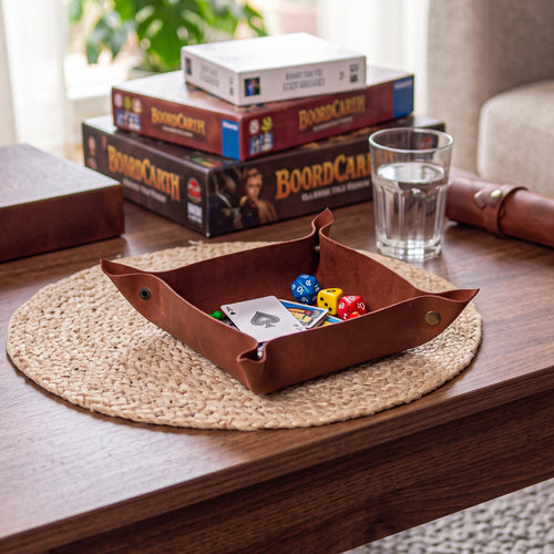 A brown leather game tray holding cards and dice, placed on a woven placemat on a wooden table. In the background, stacked BoardCARTY board games, a glass of water, and other tabletop items are visible in a real indoor setting.