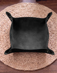A black suede-like square bowl or tray with flared corners, photographed from above on a round woven placemat over a wooden table.