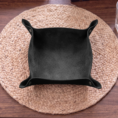 A black suede-like square bowl or tray with flared corners, photographed from above on a round woven placemat over a wooden table.