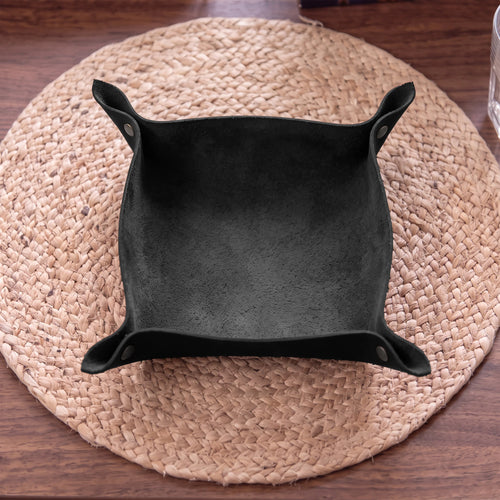 A black suede-like square bowl or tray with flared corners, photographed from above on a round woven placemat over a wooden table.