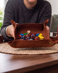 A person in a dark gray sweater holds a brown leather gaming tray filled with colorful polyhedral dice and small game pieces on a woven placemat over a wooden table.