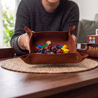 A person in a dark gray sweater holds a brown leather gaming tray filled with colorful polyhedral dice and small game pieces on a woven placemat over a wooden table.