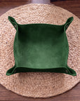 A square green suede-like tray or bowl with folded side flaps and small metal snap buttons, placed on a round woven placemat over a wooden table.