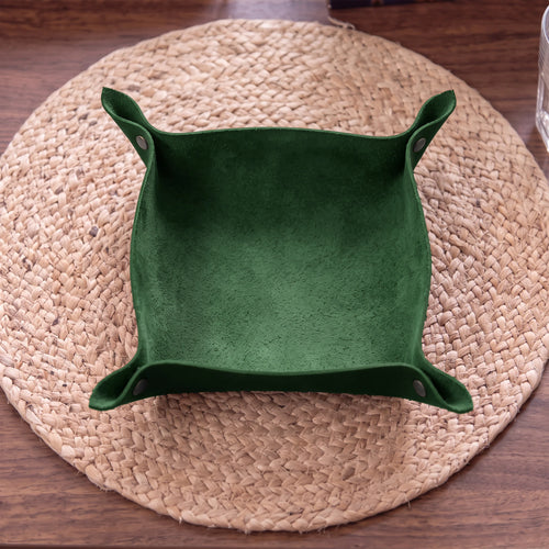 A square green suede-like tray or bowl with folded side flaps and small metal snap buttons, placed on a round woven placemat over a wooden table.