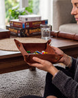 A woman holds a brown leather game tray filled with colorful game pieces on a wooden table in a cozy living room setting, with board game boxes and a glass of water in the background.