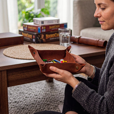 A woman holds a brown leather game tray filled with colorful game pieces on a wooden table in a cozy living room setting, with board game boxes and a glass of water in the background.