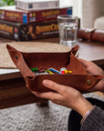 A person holds a brown leather coin purse filled with colorful game pieces on a wooden table, with board game boxes and a glass of water in the background.