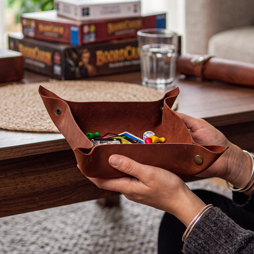A person holds a brown leather coin purse filled with colorful game pieces on a wooden table, with board game boxes and a glass of water in the background.