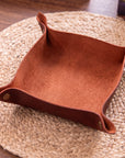 A square brown leather storage bin with flared handles and brass grommets, resting on a round woven placemat over a wooden table.