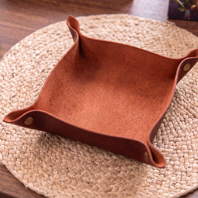 A square brown leather storage bin with flared handles and brass grommets, resting on a round woven placemat over a wooden table.