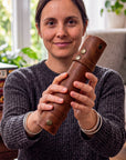 A woman holding a brown leather cylindrical case with brass snaps, shown in a cozy indoor setting with a window and plants in the background.