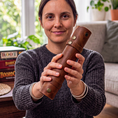 A woman holding a brown leather cylindrical case with brass snaps, shown in a cozy indoor setting with a window and plants in the background.