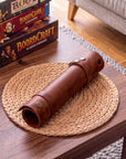 A brown leather cylindrical sleeve or roll with brass-tone snaps rests on a round woven placemat on a wooden table. In the background, stacked board game boxes sit on a sofa, and part of a chair and rug are visible, indicating a real indoor environment.