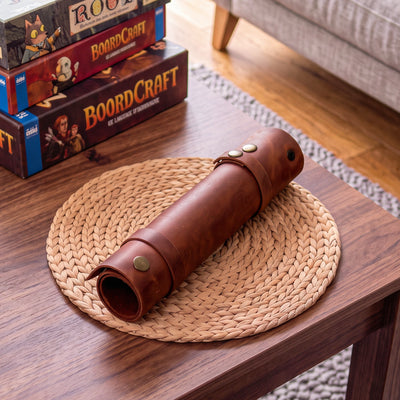 A brown leather cylindrical sleeve or roll with brass-tone snaps rests on a round woven placemat on a wooden table. In the background, stacked board game boxes sit on a sofa, and part of a chair and rug are visible, indicating a real indoor environment.