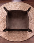 A dark brown suede-like square bowl or dish with flared sides, placed on a round woven straw placemat over a wooden table.