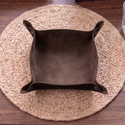 A dark brown suede-like square bowl or dish with flared sides, placed on a round woven straw placemat over a wooden table.