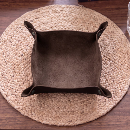 A dark brown suede-like square bowl or dish with flared sides, placed on a round woven straw placemat over a wooden table.