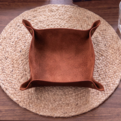 A square brown suede or faux-suede storage basket with folded side tabs, shown from above on a round woven placemat over a wooden table.