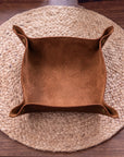 A square brown suede or faux-suede storage bin with flared sides and snap tabs, placed on a round woven placemat over a wooden table.