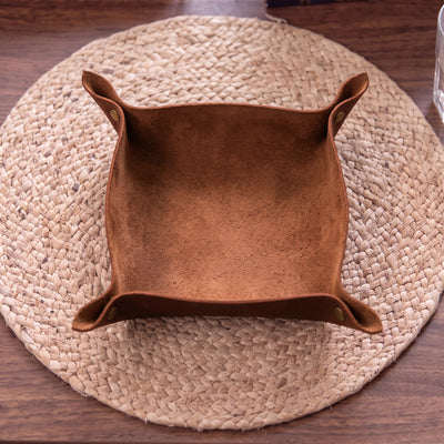 A square brown suede or faux-suede storage bin with flared sides and snap tabs, placed on a round woven placemat over a wooden table.