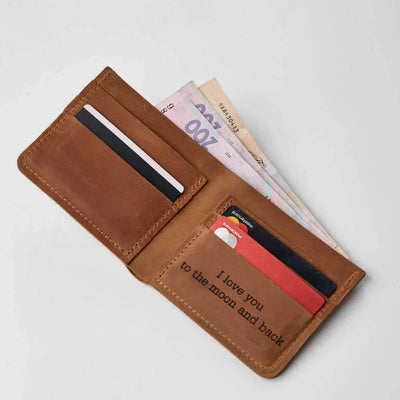 A brown leather bifold wallet shown open on a light gray background, filled with cards and cash. The inside front pocket has the printed phrase 'I love you to the moon and back' in black text.