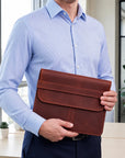 A man in a light blue button-down shirt holds a brown leather document pouch or laptop sleeve in an office setting.